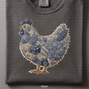 May include: A dark grey t-shirt with a patchwork chicken design in blue and cream. The chicken is made of various fabric patterns and has a stitched outline. The word "Pepper" is printed at the bottom of the shirt.