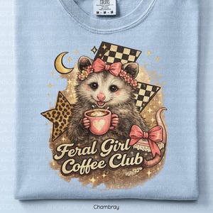 May include: Light blue t-shirt featuring a possum illustration. The possum wears a floral crown and holds a coffee cup with a heart. The text "Feral Girl Coffee Club" is below the possum. The design includes a star, moon, and checkered flag.