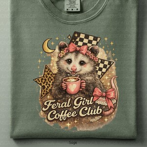 May include: Sage green t-shirt featuring a possum with a floral crown, holding a coffee cup. The design includes a crescent moon, stars, and the text "Feral Girl Coffee Club". Checkered flags and a leopard print star are also present.