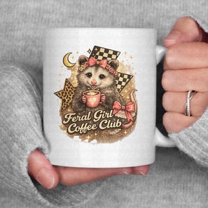May include: White ceramic mug featuring a whimsical illustration of an opossum holding a coffee cup. The opossum wears a pink bow and is surrounded by stars, a crescent moon, and the text "Feral Girl Coffee Club."