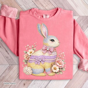 Könnte beinhalten: Ein pinkfarbenes Sweatshirt mit einem Strass-Hasen-Design. Der Hase hat eine Schleife und florale Akzente in Pastellfarben. Der Text "Watermelon" ist unten links zu sehen.