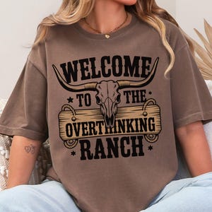 May include: A brown t-shirt with the text "WELCOME TO THE OVERTHINKING RANCH" in black. The design features a bull skull with horns above a wooden sign. The shirt is a relaxed fit.