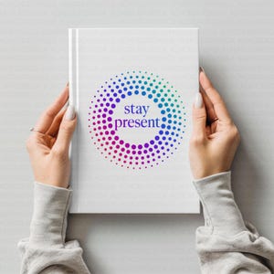 Op de afbeelding: Wit notitieboek met de woorden "stay present" in een kleurrijk stippenontwerp. Het ontwerp heeft een verloop van blauwe, groene, paarse en roze stippen. Het notitieboek wordt door twee handen vastgehouden.