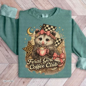 May include: Light green sweatshirt featuring a possum with a floral crown and bow, holding a coffee cup. The design includes a crescent moon, stars, and the text "Feral Girl Coffee Club". The sleeves have a cuffed design.
