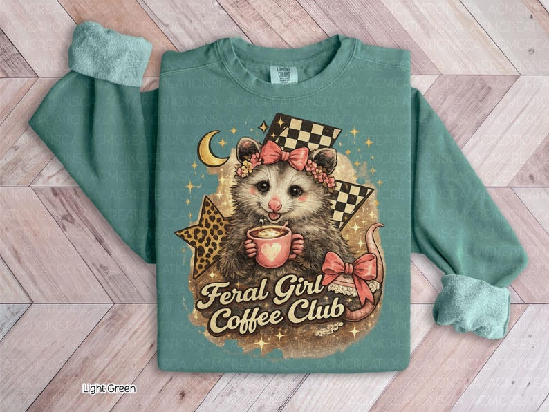 May include: Light green sweatshirt featuring a possum with a floral crown and bow, holding a coffee cup. The design includes a crescent moon, stars, and the text "Feral Girl Coffee Club". The sleeves have a cuffed design.