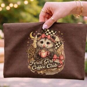 May include: A brown zippered pouch featuring a cartoon opossum holding a coffee cup, adorned with a floral crown and bow. The text "Feral Girl Coffee Club" is displayed below the opossum. The design includes a star, moon, and checkered flag.