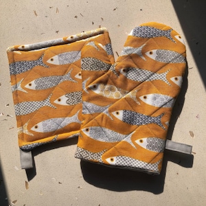 May include: A quilted oven mitt and pot holder set. The set features a repeating pattern of fish in gray, white, and yellow-orange. The mitt has a loop for hanging. The pot holder is square with a gray loop.