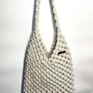 May include: An off-white macramé shoulder bag featuring a dense, knotted cord pattern and a single integrated strap. A small, rectangular dark brown label with the text MANGO TIDE is attached to the upper right side of the bag's front panel.