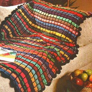 Peut inclure: Une couverture crochetée à la main, aux couleurs vives et au motif texturé en forme de grille. La couverture présente un éventail de couleurs vives, notamment le rouge, le bleu, le vert et le jaune, avec une bordure festonnée noire. La couverture est posée sur un canapé beige.
