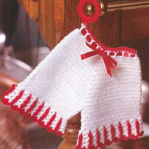 Może przedstawiać: Biała skarpetka na drutach z czerwoną lamówką i czerwoną satynową kokardką. Skarpetka wisi na metalowym haczyku.