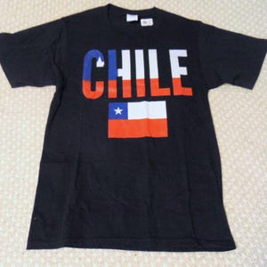 May include: Black t-shirt featuring the word "CHILE" in red, white, and blue block letters. Below the word is the flag of Chile, with a white star on a blue square, and red and white stripes.