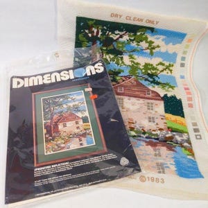 May include: A Dimensions needlepoint kit titled Springtime Reflections featuring a printed canvas of a stone house by a stream with trees and a blue sky. The kit includes a plastic bag with product text and a separate canvas with color swatches.
