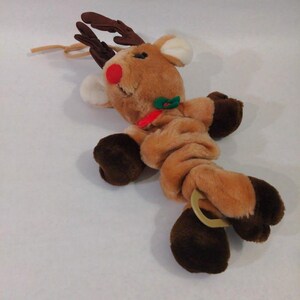 May include: A plush reindeer toy with light brown fur, dark brown hooves, and dark brown antlers. The reindeer features a bright red nose, white inner ears, and a green and red collar. A tan elastic loop is attached to the top and a plastic ring is at the base.