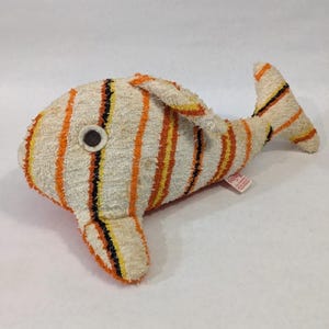 May include: A plush fish toy made of off-white terry cloth fabric featuring vertical stripes in orange, yellow, and black. The fish has a circular black and white eye and a small white fabric tag attached to its side with visible text.
