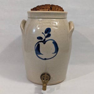 May include: A beige stoneware crock with two side handles, a cork stopper, and a brass spigot at the base. The front features a blue painted apple design and the text Walnut Hill Stoneware, Des Moines, Iowa, Living History Farms.