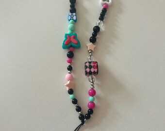 May include: A beaded phone charm featuring a variety of colorful beads. The charm includes black, pink, and turquoise beads, along with star and butterfly-shaped accents. A black cord is attached for securing the charm to a phone or other device.