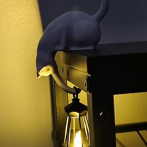 Può includere: Lampada decorativa a forma di gatto in un ambiente buio. Il gatto è appollaiato su una struttura metallica nera, con la testa e il collo curvi verso il basso. Una piccola lanterna con una calda luce è appesa sotto.