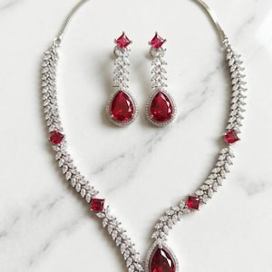 May include: A silver-toned jewelry set featuring a necklace and matching earrings. The necklace and earrings have a leaf-like design with clear, sparkling stones and vibrant red teardrop-shaped and square-cut gemstones. The set is displayed on a white surface.