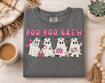 May include: A charcoal gray crewneck t-shirt featuring the text "BOO BOO CREW" in pink patterned letters above a graphic of four white ghosts dressed as medical professionals with various pink medical accessories and a pumpkin bucket.