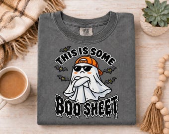 May include: A charcoal gray t-shirt featuring a graphic of a white ghost wearing an orange baseball cap and black sunglasses, with arms crossed. The text reads THIS IS SOME BOO SHEET, surrounded by small black bats on a dark gray background.