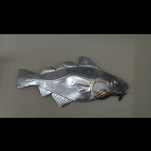 Könnte beinhalten: Metall-Wandkunst, die einen Fisch mit einem Fisch im Maul darstellt. Der Fisch ist silberfarben mit kupferfarbenen Akzenten an den Kiemen. Das Kunstwerk hat Flossen und Schuppen. Es ist an einer neutralfarbenen Wand befestigt.