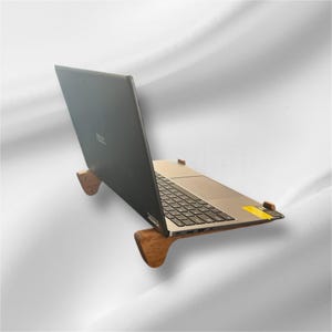 May include: A laptop computer is displayed on a wooden stand. The laptop is black and silver, with the keyboard visible. The stand is made of light brown wood and has a curved design.