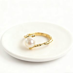 Può includere: Un anello color oro con una grande perla bianca e rotonda. L'anello ha un design a forma di bambù, con la perla tenuta da rami a forma di bambù. L'anello è presentato su un piatto bianco e rotondo.