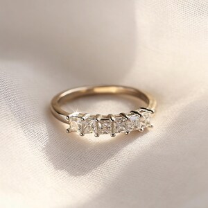 Può includere: Un anello dorato con cinque pietre preziose quadrate e trasparenti disposte in fila. L'anello è esposto su un tessuto bianco morbido, con le pietre preziose che catturano la luce e brillano. L'anello è un gioiello.