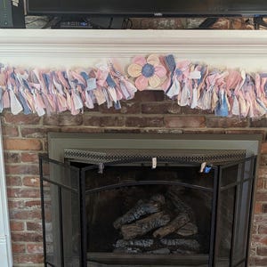 Puede incluir: Una guirnalda decorativa con tiras de tela rosa, azul y blanca cuelga sobre una chimenea de ladrillo. Una gran flor de tela está centrada en la guirnalda. La chimenea tiene una pantalla de metal negro y leños en el interior.