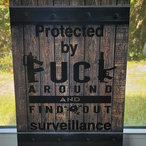 May include: A rectangular sign with a faux wood grain finish and black horizontal metal-look bands at the top and bottom. The text reads Protected by FUCK AROUND AND FIND OUT surveillance, with black rifle silhouettes integrated into the lettering.