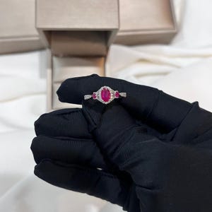 May include: A silver ring featuring an oval-cut ruby center stone, flanked by two smaller ruby stones. The ring is surrounded by a halo of small, clear gemstones. The ring is held against a black background.