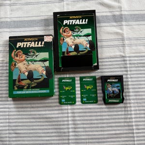 May include: A collection of vintage video game cartridges and accessories. The boxes and cartridges feature the game title "Pitfall!" with a jungle adventurer graphic. The color scheme includes green, black, and tan.