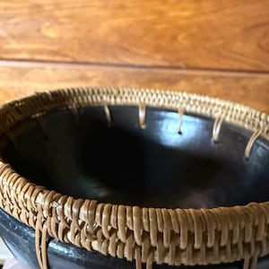 May include: A handcrafted black bowl with a woven, light brown rim. The bowl's interior is dark, contrasting with the lighter rim. The rim is made of woven natural fibers, adding a rustic touch to the design.