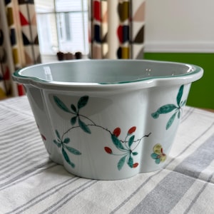 May include: A white ceramic bowl with a scalloped edge and a green trim. The bowl is decorated with hand-painted green leaves, red berries, and a yellow butterfly. The bowl is sitting on a striped tablecloth.