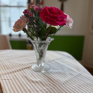 May include: A clear glass vase filled with a bouquet of fresh flowers, including a large, vibrant pink rose and several smaller pink carnations. The vase sits on a striped tablecloth, creating a simple yet elegant floral arrangement.