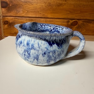 May include: A handmade ceramic bowl with a handle, featuring a blue and white speckled glaze. The bowl has a rounded shape with a pouring spout and a sturdy handle. The interior and exterior are decorated with a unique, artistic pattern.