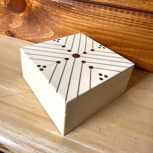 May include: A cream-colored, geometric box with a diamond-shaped lid. The lid features a pattern of black lines and brown dots radiating from a central point. The box is set against a wooden background.