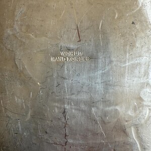 K&ouml;nnte beinhalten: Nahaufnahme einer strukturierten, silberfarbenen Metalloberfl&auml;che mit den Worten "WORLD HAND FORGED" eingepr&auml;gt. Das Metall weist Kratzer und Unvollkommenheiten auf, was ihm ein Vintage-Aussehen verleiht.