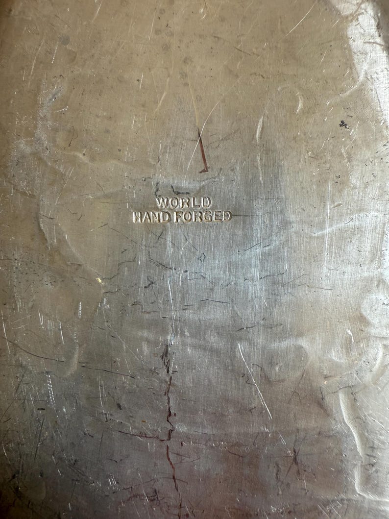 K&ouml;nnte beinhalten: Nahaufnahme einer strukturierten, silberfarbenen Metalloberfl&auml;che mit den Worten "WORLD HAND FORGED" eingepr&auml;gt. Das Metall weist Kratzer und Unvollkommenheiten auf, was ihm ein Vintage-Aussehen verleiht.