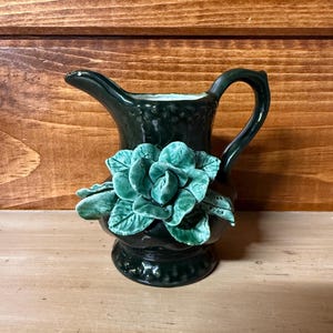 May include: A dark green ceramic pitcher with a handle and spout, adorned with a turquoise-colored floral design. The pitcher has a textured surface and sits on a wooden surface.