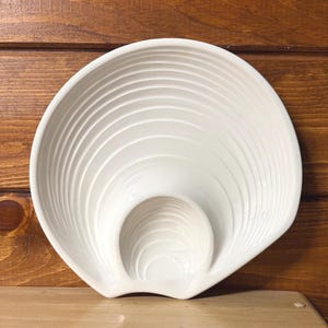 May include: A white, shell-shaped serving dish with a textured, ridged design. The dish features a central, smaller bowl area, and is made of ceramic. The dish is displayed on a wooden surface.