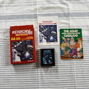 May include: A collection of Atari Asteroids video game items. Includes a red box with "Asteroids" text, a game cartridge, a manual, and a catalog. The box says "66 Video Games".