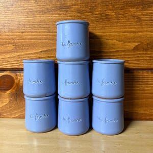 May include: A stack of seven light blue ceramic jars with the text "la fermière" embossed on the front. The jars are cylindrical with a slightly flared rim and a smooth, matte finish. The jars are arranged in a pyramid shape.