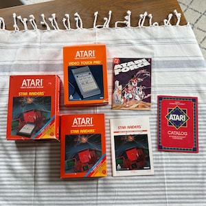 May include: A collection of vintage Atari video game cartridges and accessories. The items include boxes for "Star Raiders" and "Video Touch Pad", along with a "Star Force" comic book and a catalog. The boxes are orange and red with retro graphics.