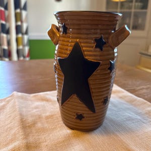 May include: A brown ceramic vase with a ribbed texture and two handles. The vase is decorated with black star shapes of varying sizes. The vase is sitting on a white cloth.