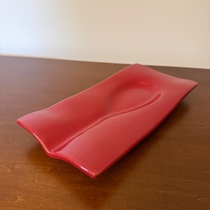 May include: A rectangular red ceramic tray with a smooth, glossy finish and a distinct, undulating wave-like surface. The tray features a central elongated depression that runs along its length, creating a gentle, curved contour across the piece.