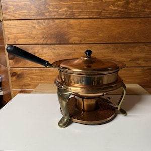 May include: A copper fondue pot with a polished finish, featuring a long black handle and a matching lid with a small black knob. The pot rests on a circular stand supported by three curved brass legs, with a central burner component underneath.