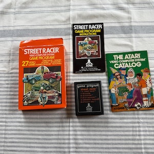 May include: An orange box and three game program booklets for the Atari video game "Street Racer." The box and booklets feature illustrations of cars and motorcycles. The Atari catalog is green.