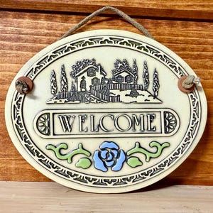 May include: Oval ceramic welcome sign with a cream background, featuring a house scene and the word "WELCOME". The sign has a decorative border, a blue rose, and green accents. It hangs from a brown rope.