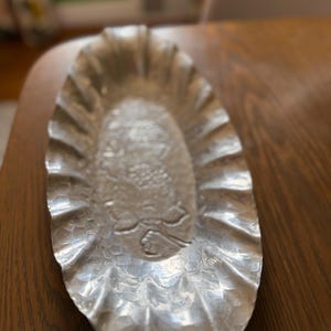 K&ouml;nnte beinhalten: Silberfarbene, ovale Servierschale mit strukturierter Oberfl&auml;che und ger&uuml;schtem Rand. Die Mitte der Schale weist ein detailliertes gepr&auml;gtes Design auf. Die Schale steht auf einem Holztisch.